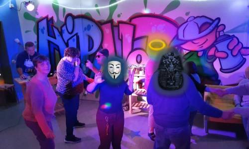 Tanzende Personen in einer Disco mit Masken. Die Wand im Hintergrund ist ein Graffiti. In der linken Ecke steht ein DJ